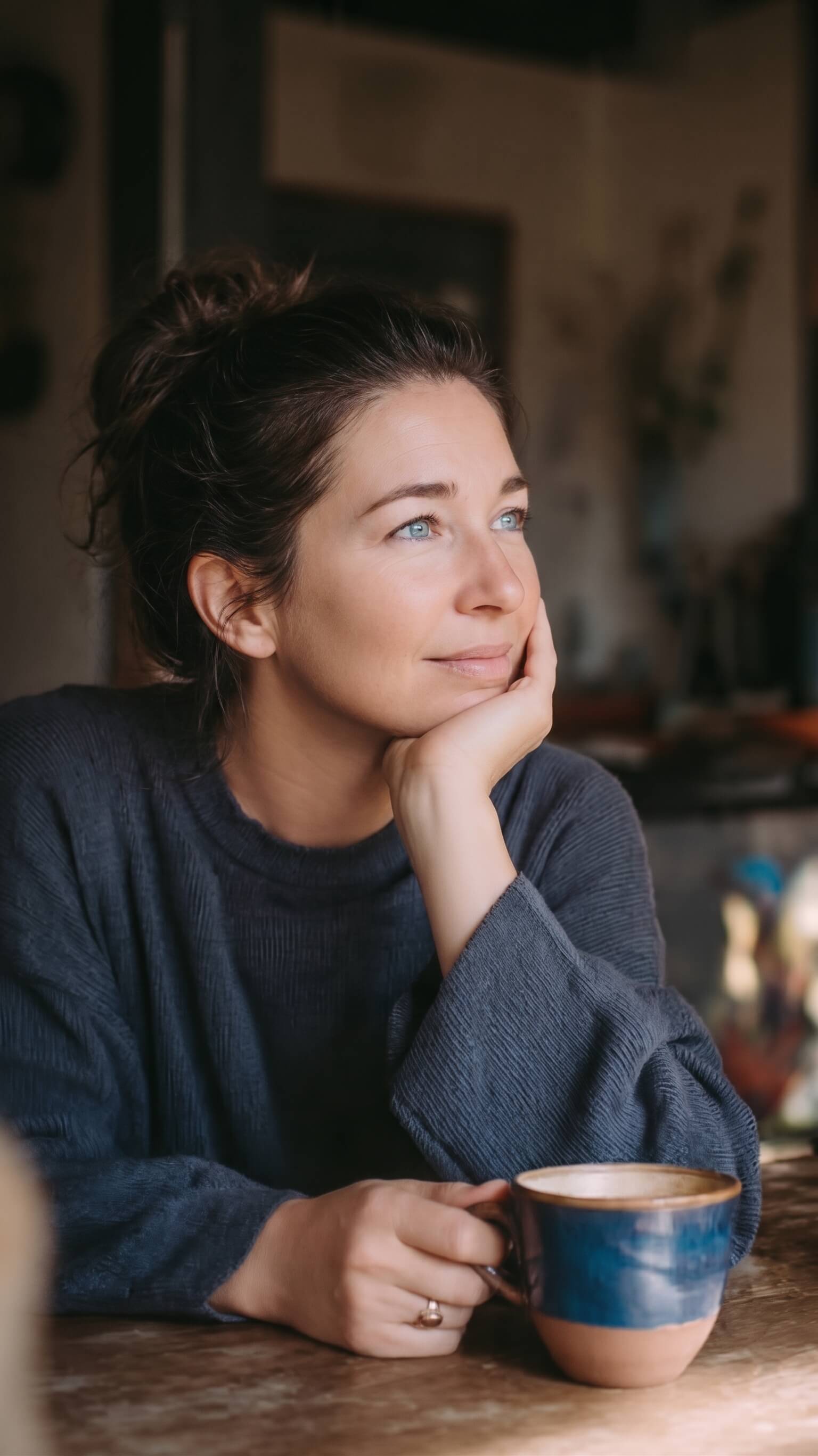 A woman with gray hair smiles thoughtfully in a warm kitchen. She wears a green knit sweater. She rests her chin on one hand and holds a red ceramic coffee mug with the other. Her relaxed posture shows a calm and recovered nervous system.